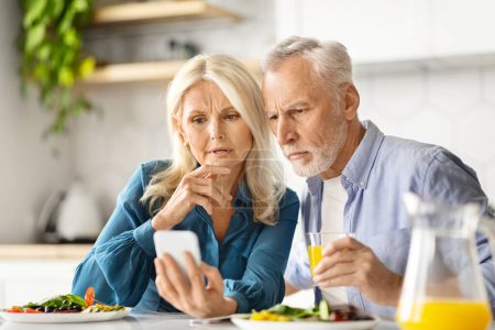 Téléchargez les photos : Conjoints aînés inquiets lisant des nouvelles sur Smartphone pendant le petit déjeuner dans la cuisine, homme et femme âgés inquiets assis à la table et regardant l'écran de l'appareil, J'ai un mauvais message, Gros plan - en image libre de droit