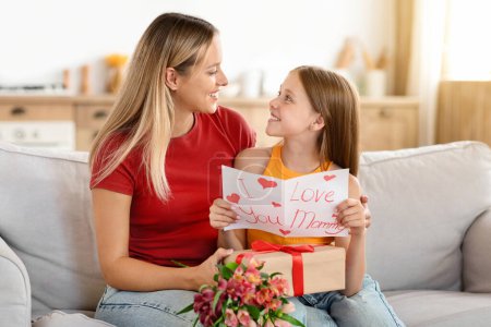 Foto de Enhorabuena. Amante hija preadolescente saludo feliz mamá con el Día de las Madres, dándole su tarjeta hecha a mano y caja de regalo, sentado en el sofá en el interior de casa, abrazo, espacio libre - Imagen libre de derechos