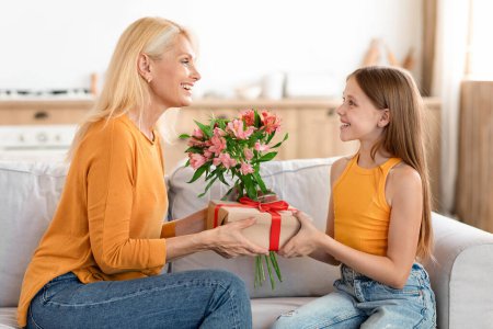 Photo for Congratulations. Loving preteen girl granddaughter greeting happy granny with birthday, giving her flowers and gift box, sitting on sofa at home interior, smiling to each other, free space - Royalty Free Image