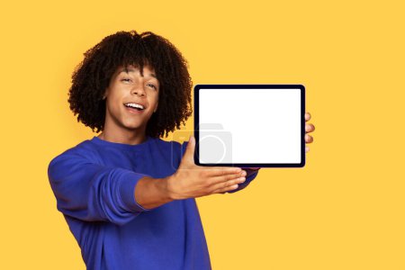 Foto de Oferta en línea. Hombre negro guapo mostrando tableta digital con pantalla blanca, joven afroamericano que demuestra espacio libre de la copia para el diseño del anuncio, de pie sobre fondo amarillo, burla - Imagen libre de derechos