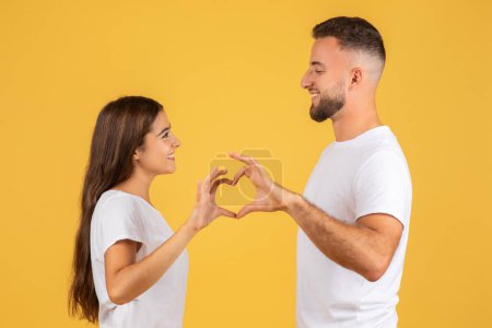 Photo for Satisfied millennial european wife and husband, making heart gesture with hands, enjoy tender moment, love, isolated on yellow studio background, profile. Date and romantic lifestyle - Royalty Free Image