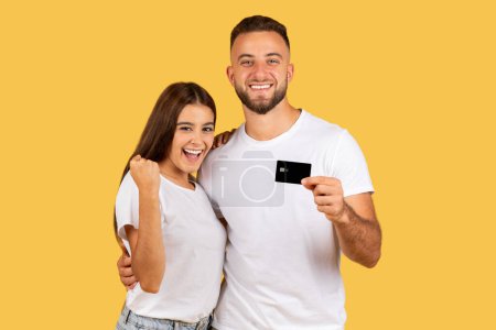 Photo for Glad young european family in white t-shirts, with credit card hugs, celebrate success, win, isolated on yellow studio background. Ad and offer, win, sale recommendation, shopping, cashback - Royalty Free Image