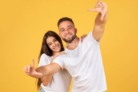 Photo for Glad young european man and woman in white t-shirts making frame with hands, capturing a moment of happiness, isolated on yellow studio background. Emotions, create, fun and lifestyle - Royalty Free Image