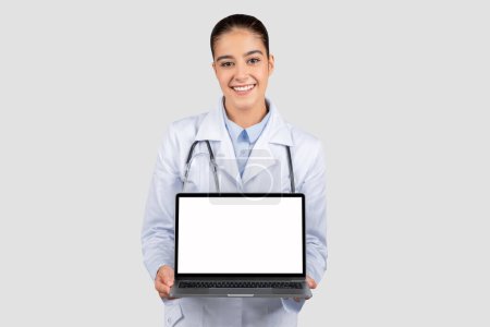 Téléchargez les photos : Joyeux caucasien jeune femme médecin en manteau blanc montrer ordinateur portable avec écran blanc, isolé sur fond de studio gris. Application de soins médicaux, recommandation du site Web - en image libre de droit
