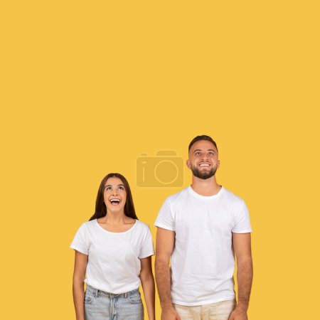 Photo for Happy shocked millennial european couple in white t-shirts, look up at empty space, isolated on yellow studio background. Ad and offer, sale and lifestyle, surprise emotion - Royalty Free Image