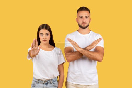Photo for Angry strict millennial european man and lady in white t-shirts make stop gesture with hand, crossed arms, isolated on yellow studio background. End, fight with harassment, rejection - Royalty Free Image
