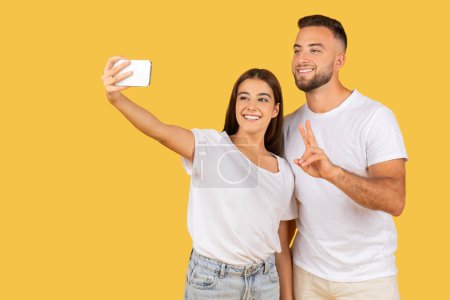 Photo for Happy millennial caucasian couple in white t-shirts have fun, enjoy date, show peace sign, make selfie on smartphone, isolated on yellow studio background. Relationships, photo for blog, video call - Royalty Free Image
