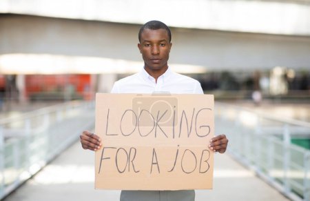 Téléchargez les photos : Homme noir sans emploi portant une chemise portant une pancarte avec la recherche d'une affiche de texte de travail, contrarié homme afro-américain malheureux a perdu son travail, souffrant de la crise du chômage, posant à l'extérieur, copier l'espace - en image libre de droit