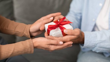 Photo for Closeup of young couples hands holding wrapped gift box sitting on sofa indoor, symbolizing romantic presents exchange, love and celebration on anniversary or Valentines day. Cropped, panorama - Royalty Free Image