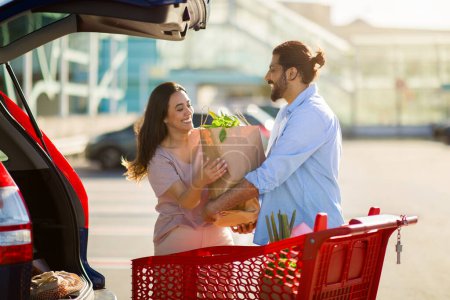 Téléchargez les photos : Heureux couple moyen-oriental riant et partageant un moment tout en tenant un sac d'épicerie entre eux par le coffre de leur voiture dans un parking ensoleillé - en image libre de droit