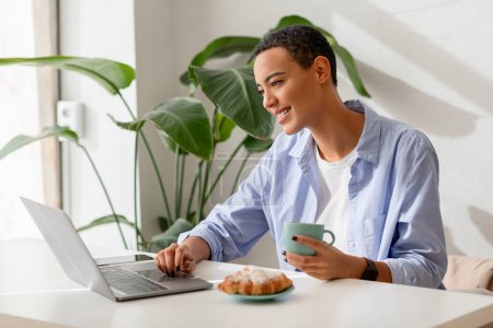 Photo for Smiling young latin lady enjoy cup of coffee and croissant at table and video call on laptop, chat in cafe, office interior. Communication remotely, app for business, work - Royalty Free Image