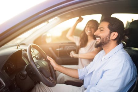 Téléchargez les photos : Joyeux couple arabe avec chauffeur et femme dansant, tous deux profitant d'une balade en voiture par une journée ensoleillée avec des rayons de soleil rayonnant dans - en image libre de droit