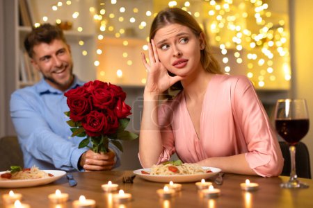 Photo for Woman appears unconvinced as smiling man offers bouquet of red roses during candlelit dinner with a hint of romantic tension - Royalty Free Image