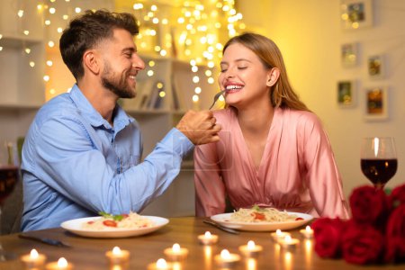 Photo for Laughing man tenderly offers fork of spaghetti to his partner during a candlelit dinner, surrounded by a bokeh of twinkling lights on background - Royalty Free Image