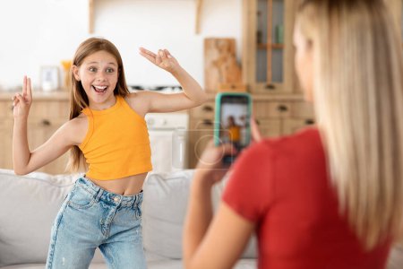 Photo for Back view of blonde mother shooting on smartphone her dancing daughter, home interior. Funny teenager girl asking mom to record video for social media, dancing and grimacing - Royalty Free Image