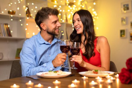 Téléchargez les photos : Couple joyeux partageant un toast avec des verres à vin rouge, profitant d'un dîner romantique sur fond de lumières douces et lumineuses - en image libre de droit