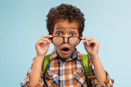 Téléchargez les photos : Étonné jeune garçon levant ses lunettes dans l'incrédulité, vêtu de chemise à carreaux colorés avec sac à dos vert, sur fond bleu apaisant, exprimant merveille - en image libre de droit
