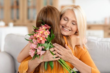 Téléchargez les photos : Portrait d'une préadolescente aimante saluant la mamie heureuse avec la Journée internationale de la femme, lui donnant des fleurs, assise sur un canapé à l'intérieur de la maison, embrassant, espace libre - en image libre de droit