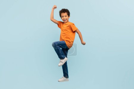 Téléchargez les photos : Jeune garçon exubérant en t-shirt orange célébrant avec poing levé et genou, mettant en valeur l'énergie et le succès, sur fond bleu doux, symbolisant le triomphe - en image libre de droit