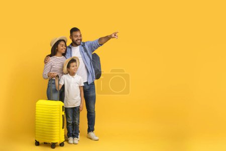 Foto de Concepto de Viaje Familiar. Padres afroamericanos felices con su hijo pequeño llevando maletas y señalando a un lado el espacio de copia en el fondo amarillo, mamá, papá y el niño listo para las vacaciones, panorama - Imagen libre de derechos