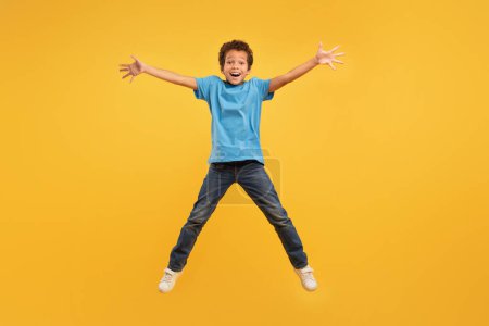 Photo for Ebullient boy mid-jump with arms outstretched, wearing light blue t-shirt and jeans, captured against vibrant yellow background, embodying freedom and happiness - Royalty Free Image
