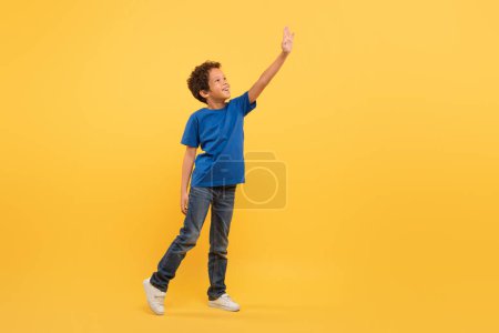 Foto de Niño estudiante negro con expresión alegre, estirando un brazo para tocar algo invisible, vestido con camiseta azul, sobre fondo amarillo brillante, lleno de imaginación - Imagen libre de derechos