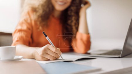 Photo for Curly-haired woman in a focused state writing in a notebook with a pen, a cup of coffee and laptop on the desk in a bright workspace, closeup, panorama - Royalty Free Image