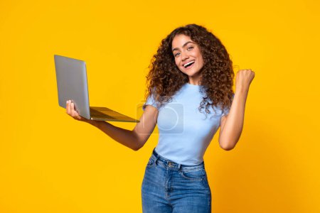 Photo for Exultant young woman with curly hair holds an open laptop in one hand and fist pumps with the other, exuding success against a yellow background - Royalty Free Image