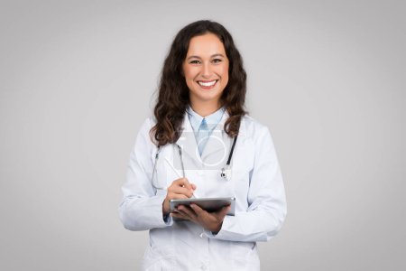 Téléchargez les photos : Femme médecin praticienne positive en manteau blanc utilisant une tablette numérique et souriante, isolée sur fond gris, prise de vue en studio. Soins de santé à distance, service de médecine app, examen - en image libre de droit