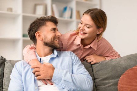 Téléchargez les photos : Les jeunes mariés partageant une étreinte décontractée sur leur canapé, se souriant, profitant d'un moment d'affection et de bonheur dans le confort de leur salon moderne à la maison. Relation amoureuse - en image libre de droit