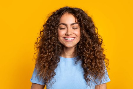 Foto de Joven contenta con los ojos cerrados y una sonrisa radiante, mostrando su pelo rizado, en una pose relajada sobre un fondo amarillo brillante - Imagen libre de derechos