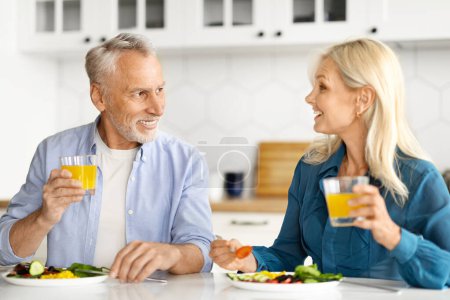Téléchargez les photos : Heureux conjoints aînés bavardant et buvant du jus d'orange pendant le petit déjeuner dans la cuisine, souriant vieil homme et femme mangeant des aliments savoureux ensemble à la maison, assis à la table et se regardant - en image libre de droit