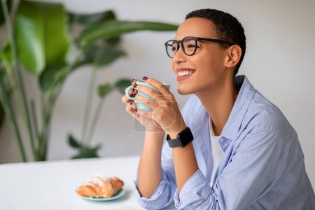 Photo for Happy calm young latin woman in glasses enjoy peace and lunch, drink cup of coffee, eat croissant, look at empty space in modern cafe, office interior. Spare time, relax, breakfast - Royalty Free Image