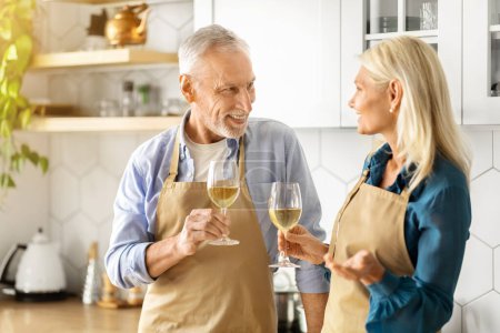 Téléchargez les photos : Couple sénior romantique buvant du vin blanc ensemble dans la cuisine, heureux conjoints plus âgés portant des tabliers tenant des verres avec boisson et souriant les uns aux autres, célébrer l'anniversaire à la maison - en image libre de droit