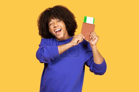 Téléchargez les photos : Joyeux jeune homme noir tenant passeport et cartes d'embarquement, heureux homme afro-américain joyeux excité pour les prochains voyages, debout isolé sur fond de studio jaune, espace de copie - en image libre de droit