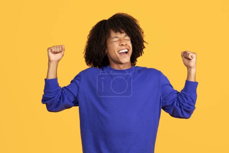 Foto de Emocional joven negro chico celebrando el éxito en amarillo fondo del estudio, lleno de alegría afroamericano masculino sacudiendo puños, guapo chico animando suerte y compartir buenas emociones, copiar espacio - Imagen libre de derechos