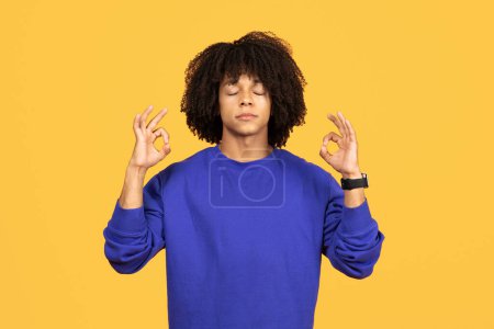 Foto de Hombre negro sereno en sudadera azul meditando con los ojos cerrados, chico afroamericano tranquilo haciendo signo ok con los dedos mientras está de pie aislado en el fondo amarillo, encarnando mindfulness, espacio de copia - Imagen libre de derechos