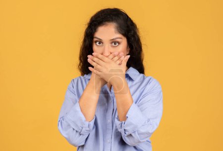 Photo for Concerned young woman with dark curly hair covers her mouth with her hands, her eyes conveying worry or a desire to keep silent, against a plain yellow background - Royalty Free Image