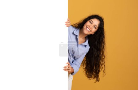 Téléchargez les photos : Joyeux latin millénaire femme brune aux cheveux longs avec grande bannière avec espace vide, recommander la vente, l'annonce et l'offre, isolé sur fond de studio orange. Attention bonnes nouvelles, soins de beauté - en image libre de droit