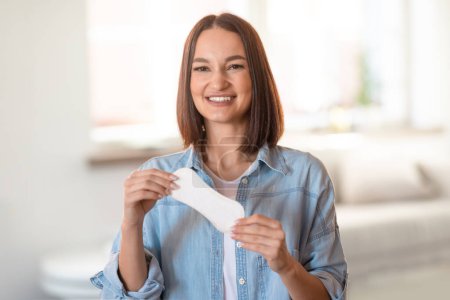 Photo for Happy young woman holds daily cotton pad and smiling to camera, posing at home interior. Woman caring for her comfort and freshness, choosing best female sanitary products - Royalty Free Image