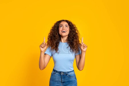Foto de Mujer alegre de pelo rizado con una camisa azul apunta hacia arriba con ambas manos, expresando felicidad y emoción, aislada sobre un vibrante fondo amarillo - Imagen libre de derechos