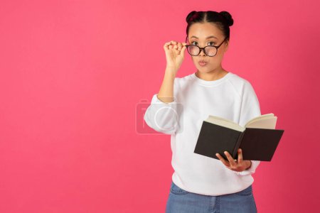 Photo for Nerdy young asian woman holding book and adjusting glasses, amazed korean female student reacting to interesting novel story, standing isolated on pink studio background, copy space - Royalty Free Image