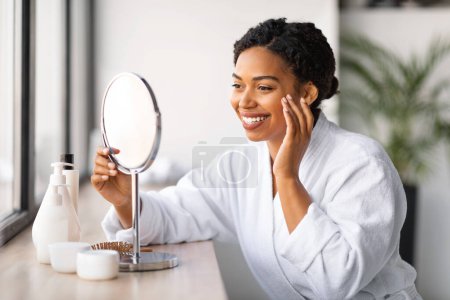 Téléchargez les photos : Belle jeune dame noire regardant dans le miroir grossissant et rendant les soins de la peau de routine à la maison, heureuse femme afro-américaine souriante en peignoir appliquant sous la crème pour les yeux, bénéficiant de soins de soi, gros plan - en image libre de droit