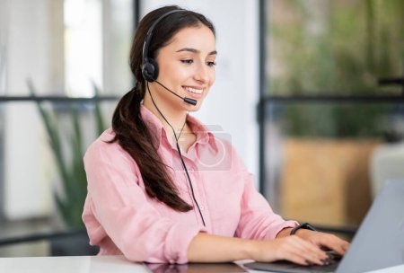 Photo for Positive pretty young european woman manager in headset typing on laptop, enjoy work in coworking office. Project, planning startup, business support, job service remotely - Royalty Free Image