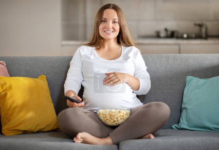 Foto de Mujer embarazada feliz relajándose en el sofá en casa, viendo la televisión y comiendo palomitas de maíz, sonriendo embarazada sosteniendo el mando a distancia y cambiando canales, disfrutando del ocio doméstico, espacio de copia - Imagen libre de derechos