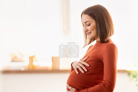 Téléchargez les photos : Grossesse et maternité. Jeune femme enceinte heureuse étreignant et touchant son ventre en pleine croissance à l'intérieur, vue de côté prise de vue de la future maman attendant bébé se sentant enfant se déplace. Espace libre pour le texte - en image libre de droit