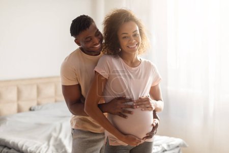 Téléchargez les photos : Jeune couple de femmes enceintes afro-américaines partage moment d'amour, mari serrant tendrement son ventre de femmes enceintes debout dans l'intérieur de la chambre moderne. Attentes et joie de la parentalité future - en image libre de droit
