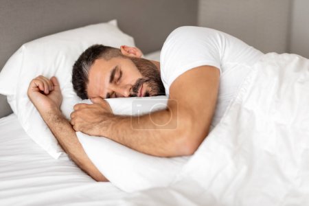 Photo for Man comfortably sleeping in bed wrapped in blanket, hugging white pillow in restful slumber and relaxation in modern bedroom setting. Shot of guy enjoying his nap and recreation - Royalty Free Image