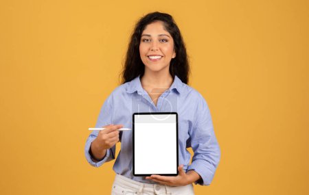 Foto de Feliz latina milenaria morena mostrar tableta con pantalla vacía y pluma, aislado en fondo de estudio naranja. Anuncio y oferta, trabajo, aplicación de estudio, recomendación del sitio web, presentación - Imagen libre de derechos