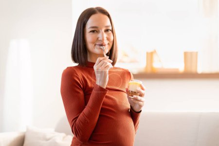 Téléchargez les photos : Régime alimentaire pendant la grossesse. Enchantée Femme enceinte tenant joyeusement un dessert savoureux, ayant une collation sucrée à la maison, incarnant la joie et les défis de la nutrition de la grossesse, souriant à la caméra - en image libre de droit